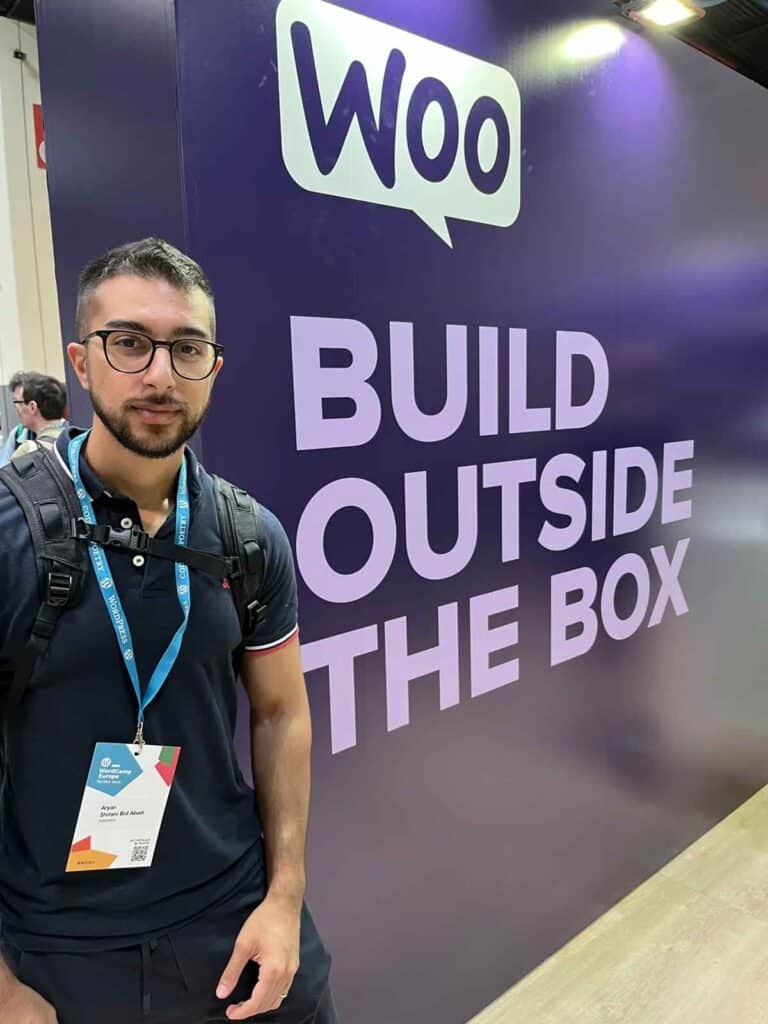 Un uomo con occhiali e zaino posa davanti al grande stand di WooCommerce con il messaggio "Build Outside The Box".